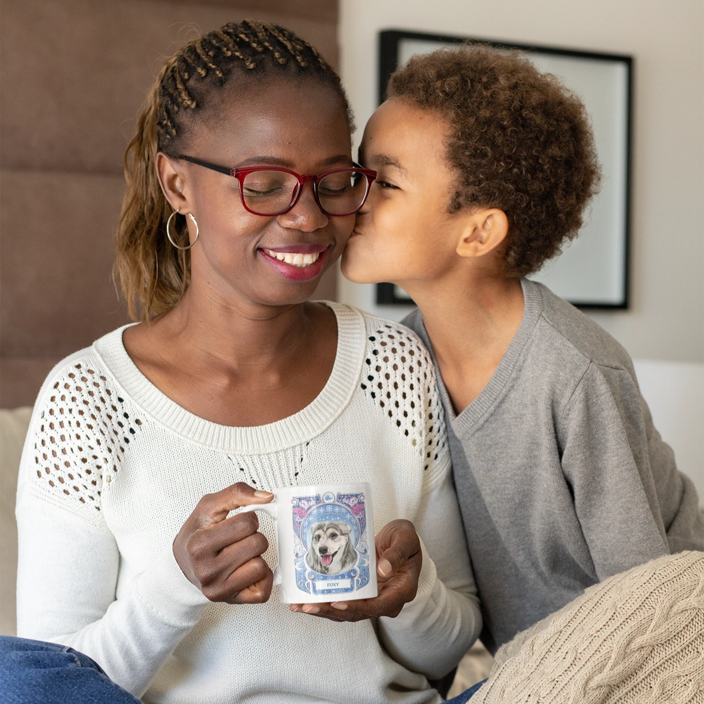 Woman holding a mug with a child kissing her on the cheek, both sitting on a couch.