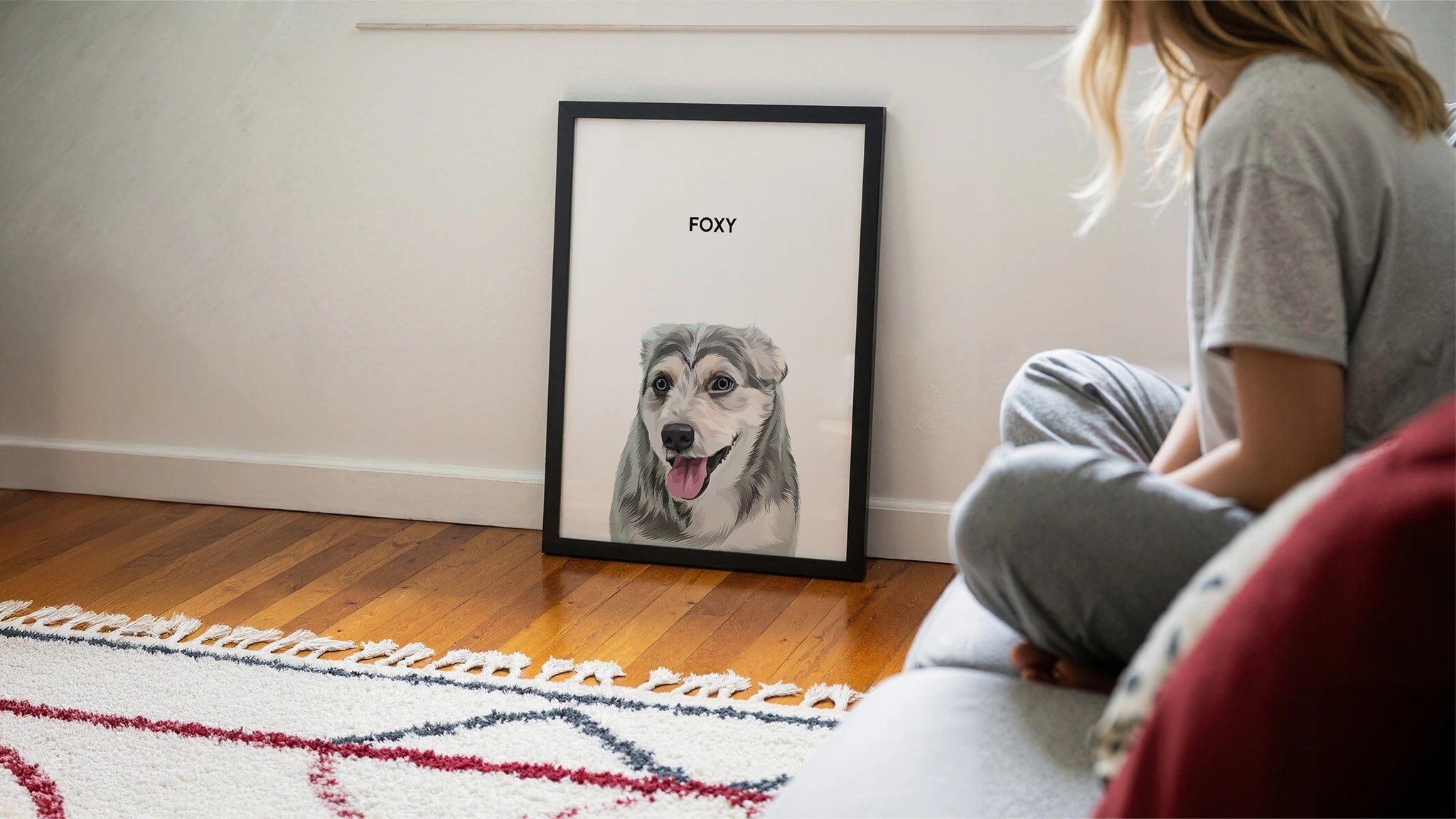 Person sitting on a couch next to a framed picture of a dog named 'Foxy' on a wooden floor.