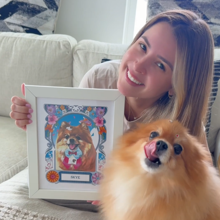 Woman holding a framed picture of a dog with a small dog sitting next to her on a couch.