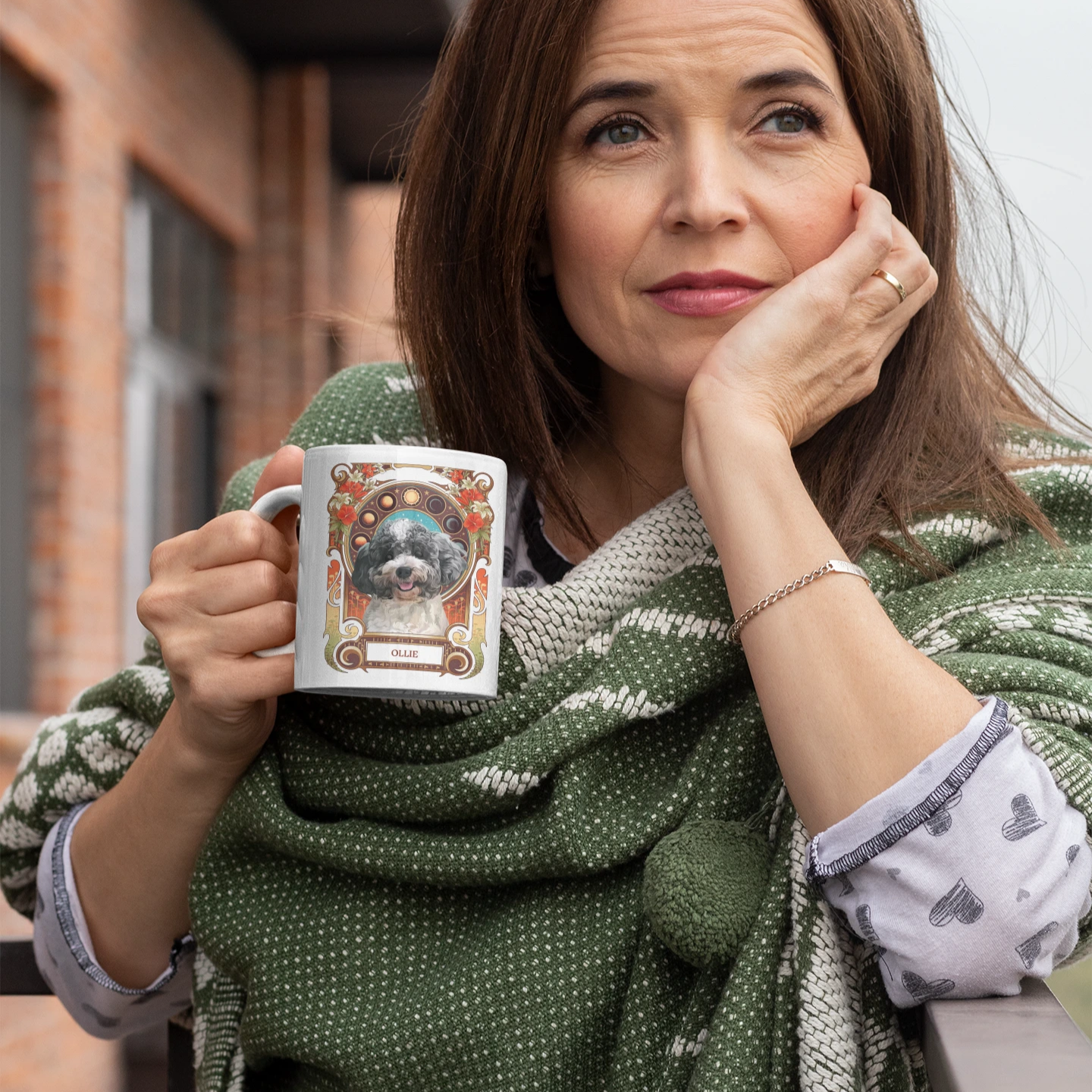 Woman holding a mug with a dog design, wrapped in a green scarf, standing outdoors.